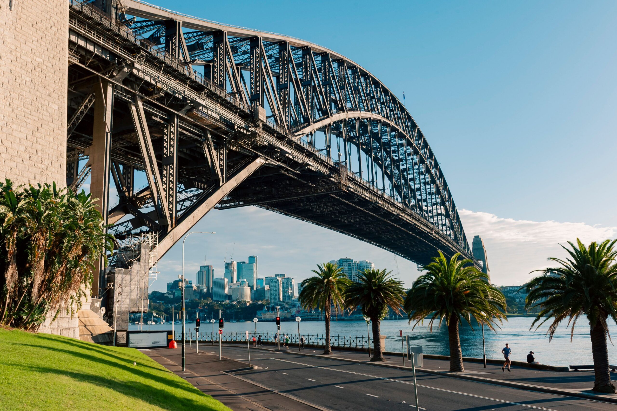 A scenic view of Sydney Harbour Bridge with clear blue sky and palm trees along a quiet road.